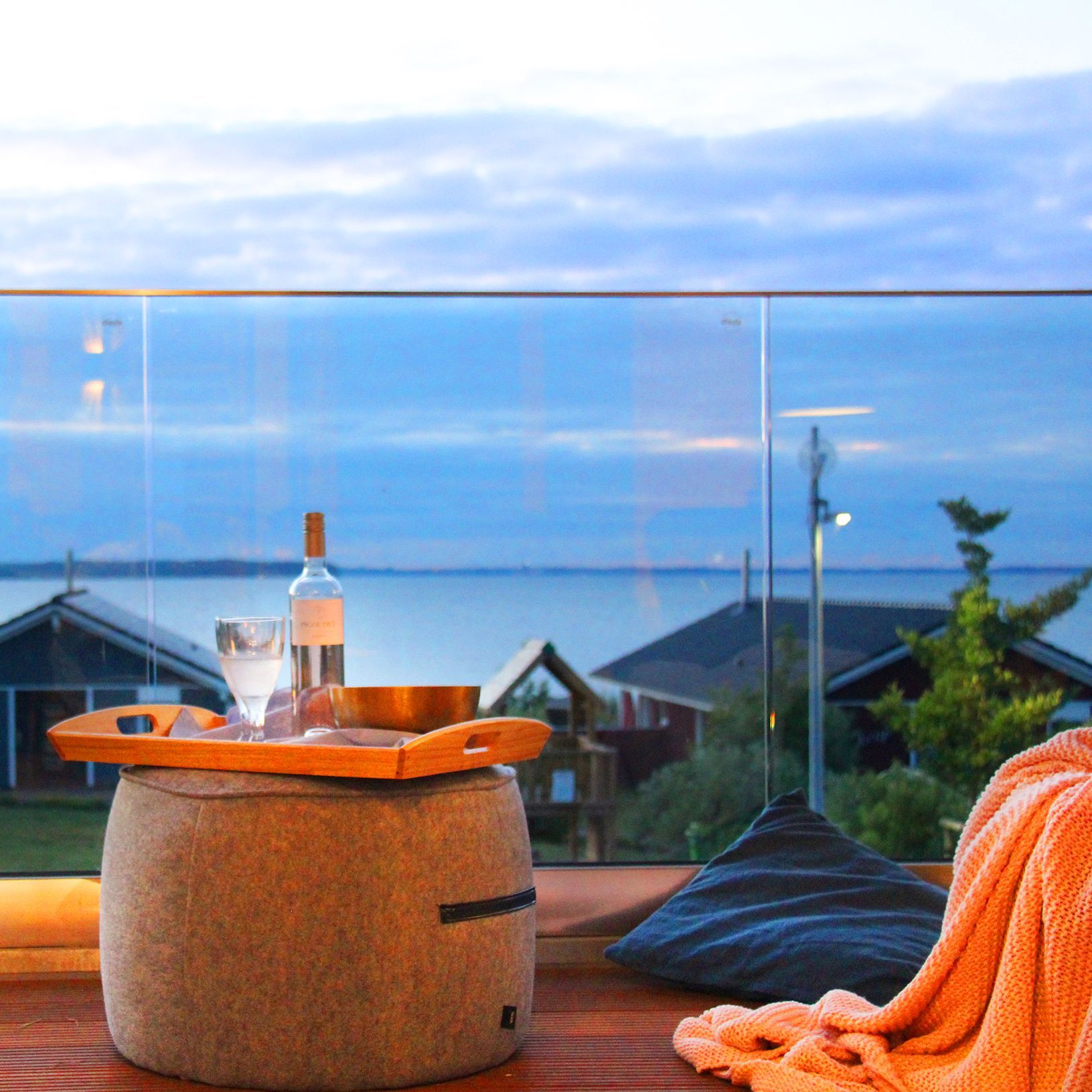 Rechts Kissen und Sandsack mit Decke, links Tablett mit Wein und Glas auf Hocker, Hintergrund Glasgeländer und Meerblick