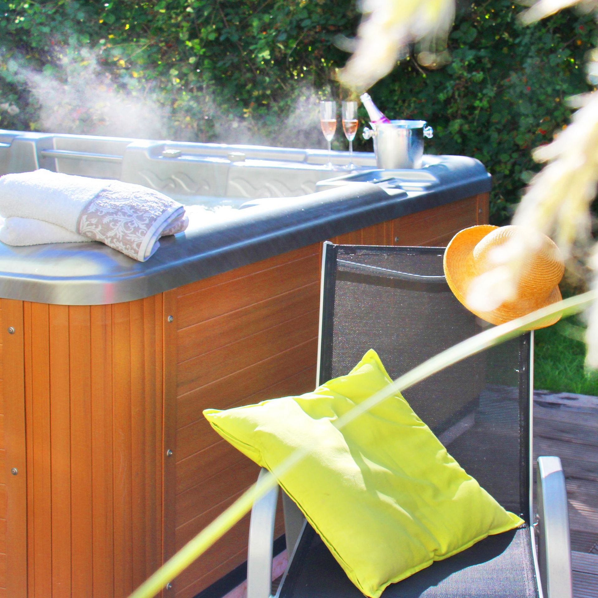 links Outdoor-Jacuzzi mit aufsteigendem Dampf, mitte ein Gartenstuhl mit Kissen, rechts blühende Gräser